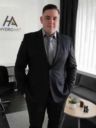 A képen egy fiatal férfi látható, aki sötét öltönyt és világos inget visel. Az Öskü városában található HydroArt Kft. épületgépészeti irodájában készült a felvétel, ahol a háttérben a cég logója és egy világos függöny látható. A férfi magabiztosan áll, kezét zsebébe dugva, ami a szakmai hozzáértést és megbízhatóságot sugallja az épületgépészet területén.