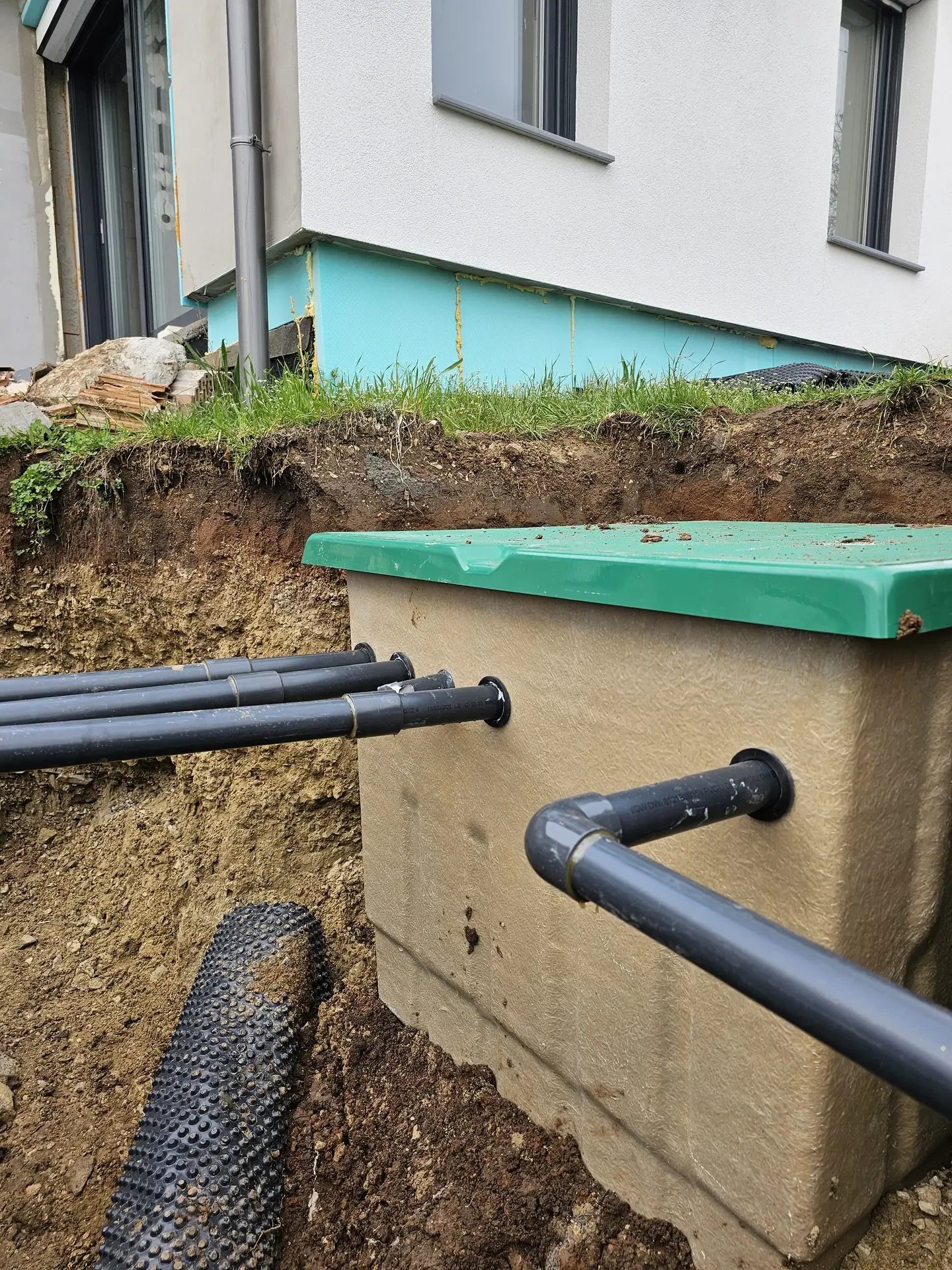 Az Ösküben található HydroArt Kft. Épületgépészet területén végzett munkáját bemutató képen az épület alapja mellett látható egy szürke betonkocka, amely vízelvezető rendszer része. A földbe fektetett fekete csövek a víz elvezetésére szolgálnak, a zöld színű fedőlap pedig a rendszer védelmét biztosítja. A háttérben az épület falának részletei is láthatók.