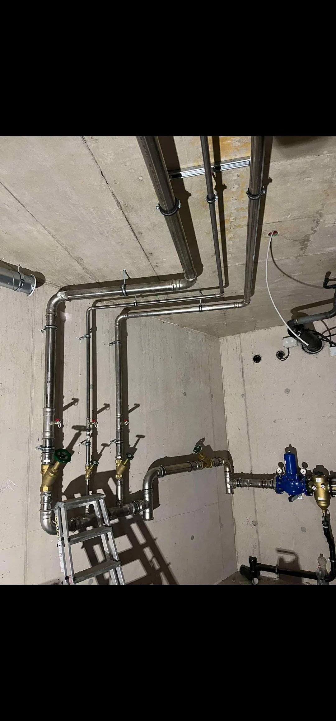 Az Ösküben található HydroArt Kft. épületgépészeti munkálatainak bemutatása látható a képen, ahol rozsdamentes acélcsövek és szelepek precíz elrendezése figyelhető meg. A csővezetékek a falon és a mennyezeten futnak, míg az alján lévő létra egyértelműsíti a munka folyamatát. Az épületgépészet szakmai színvonala tükröződik a részletek precizitásában.