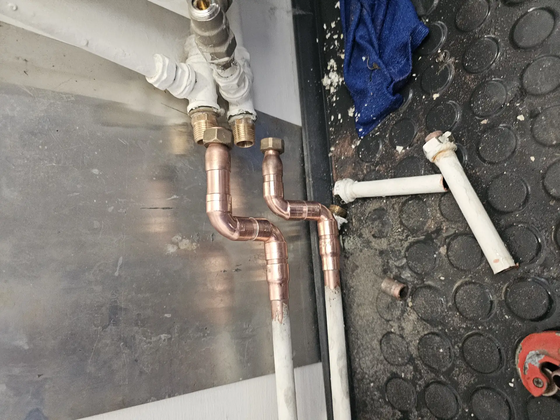 Az Öskü városában található HydroArt Kft. Épületgépészet területén végzett munkáját bemutató képen látható egy fém és műanyag csőcsatlakozás. A képen réz és fehér PVC csövek találhatók, amelyek precíz illesztésekkel vannak összekötve. A háttérben a szerelési környezet, valamint néhány szerszám és munkaeszköz is látható, ami a szakmai tevékenységre utal.