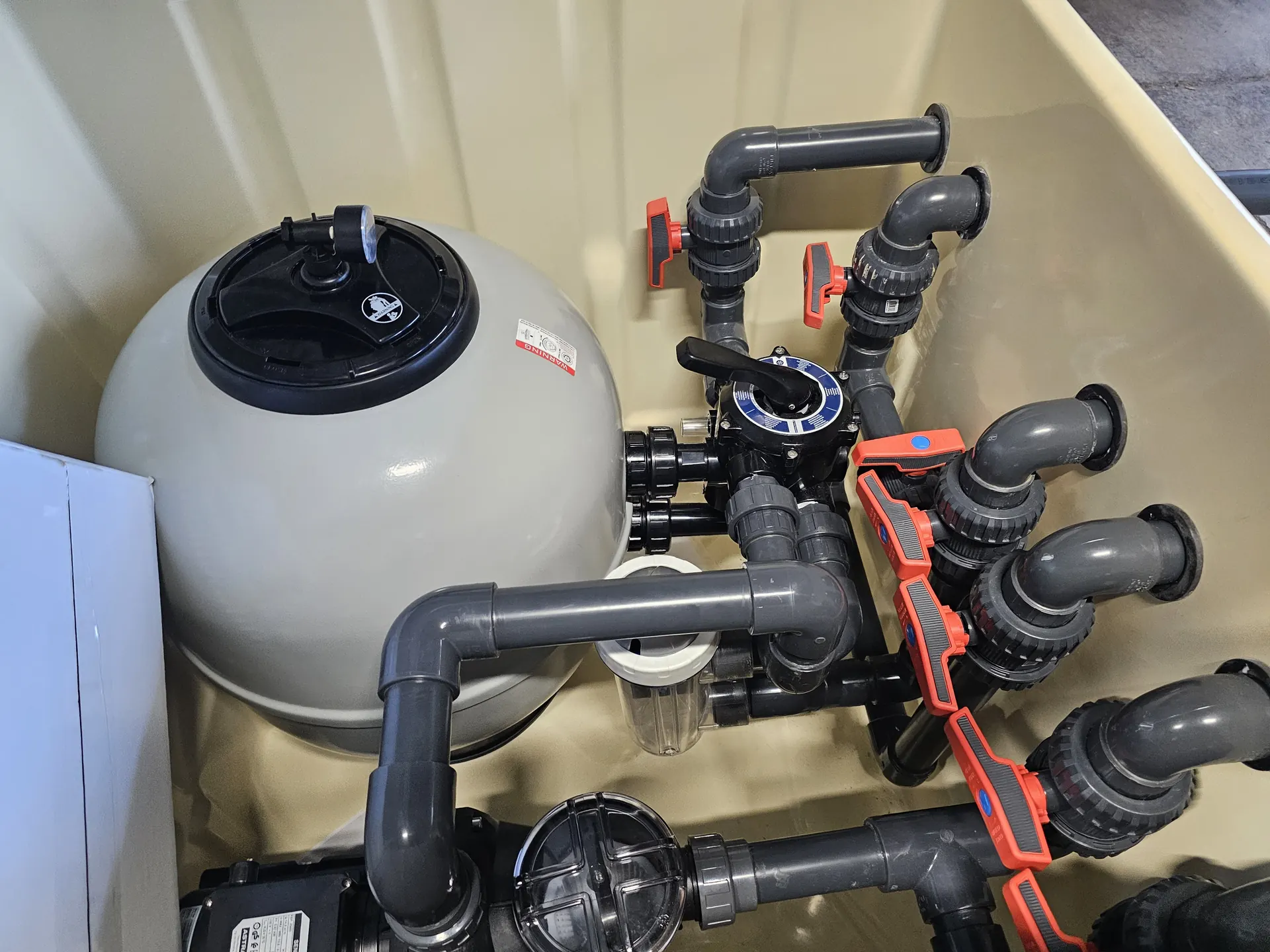 A képen a HydroArt Kft. épületgépészeti berendezései láthatók, amelyek Öskün találhatók. A tartályban egy szürke szűrő és különféle műanyag csövek, valamint szelepek láthatók, amelyek a víz áramlását szabályozzák. A precíziós kivitelezés és a modern technológia jól tükrözi az épületgépészet területén végzett munkájukat.