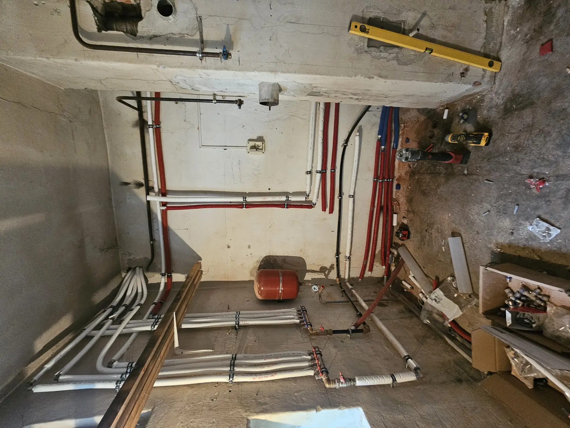 Az Öskü városában található HydroArt Kft. épületgépészeti projektje látható, amely az épület belső víz- és fűtési rendszereinek modernizálását célozza. A falon különböző színű csövek és szerelvények találhatók, a piros és fehér csövek az új rendszer részleteit mutatják. A területen szerszámok és építkezési anyagok is láthatók, amelyek az épületgépészet munkálatainak előkészítését szolgálják.