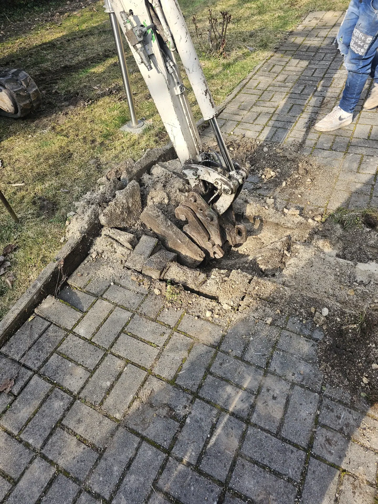 Az Ösküben található HydroArt Kft. Épületgépészet területén végzett munkáját mutatja be az image slot: gallery_06. A képen egy gép látható, amely éppen egy térkőből készült járdát bont le, a környezetben fű és föld látható. A munka során a gép a járda köveit és az alatta lévő anyagokat távolítja el.