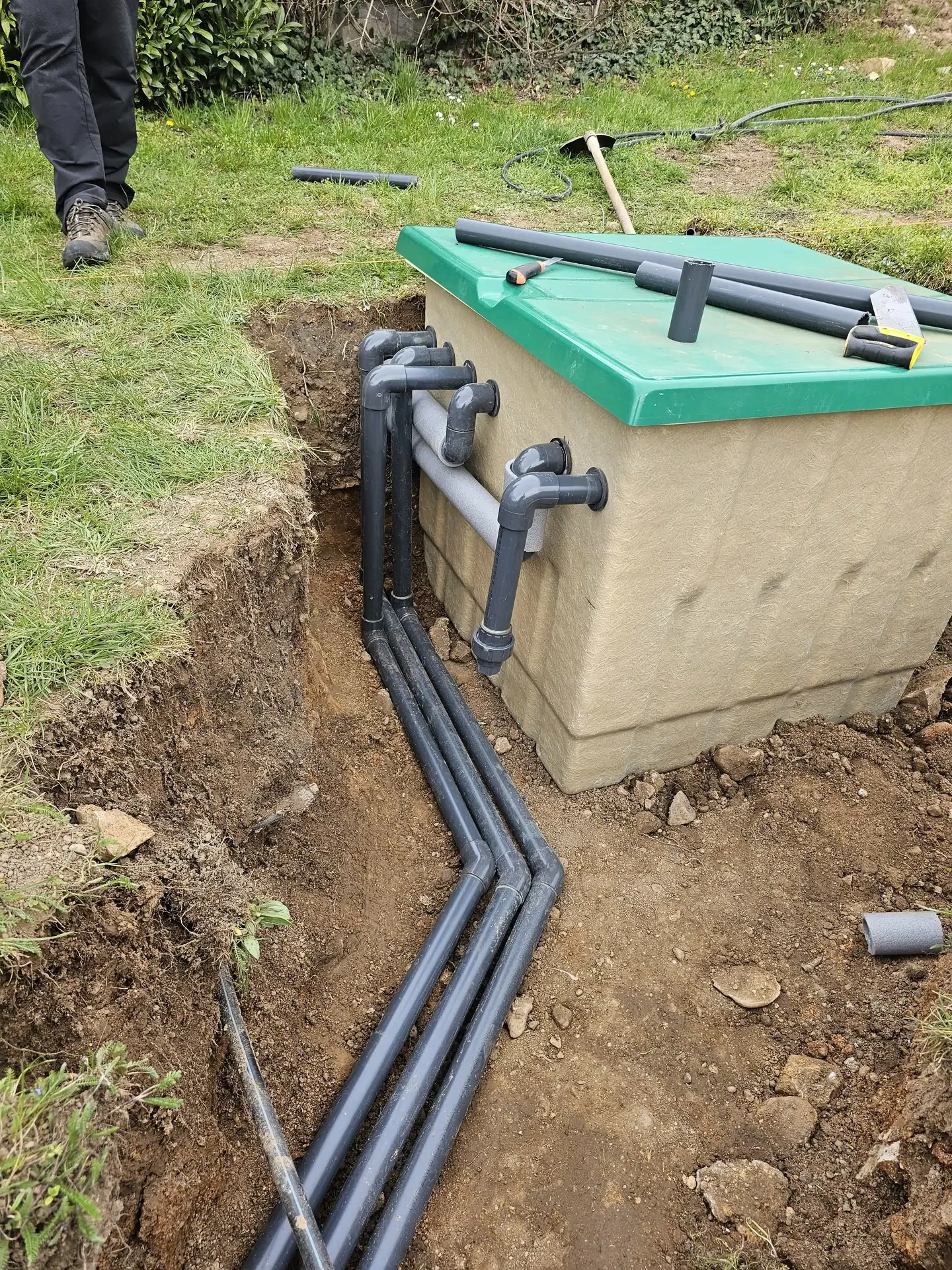 A képen egy épületgépészeti munkálat látható, ahol a HydroArt Kft. csapata Öskü városában csöveket telepít. A földbe ásott csövek mellett egy zöld fedelű tartály található, amely a víz- vagy csatornarendszer része lehet. A környező terület zöld fűvel borított, ami a munkálatok előkészítését jelzi.