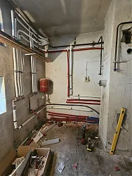 Az Ösküben található HydroArt Kft. Épületgépészet tevékenységéhez kapcsolódó képen egy építkezési terület látható, ahol a falon különböző csövek és vezetékek futnak, piros és fehér színekben. A padló borítása rendezetlen, építőanyagok és szerszámok hevernek szanaszét. A falon egy vízcsap is található, amely a gépészeti munkálatok része.