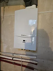 A képen egy modern, fehér színű fűtőberendezés látható, amely a HydroArt Kft. épületgépészeti szakértelmét tükrözi. A készülék a falra szerelve található, és a csövek is jól láthatóak az alján, amelyek az épületgépészet szerves részét képezik. Az Öskü városában végzett munka minőségét mutatja be ez a professzionális installáció.