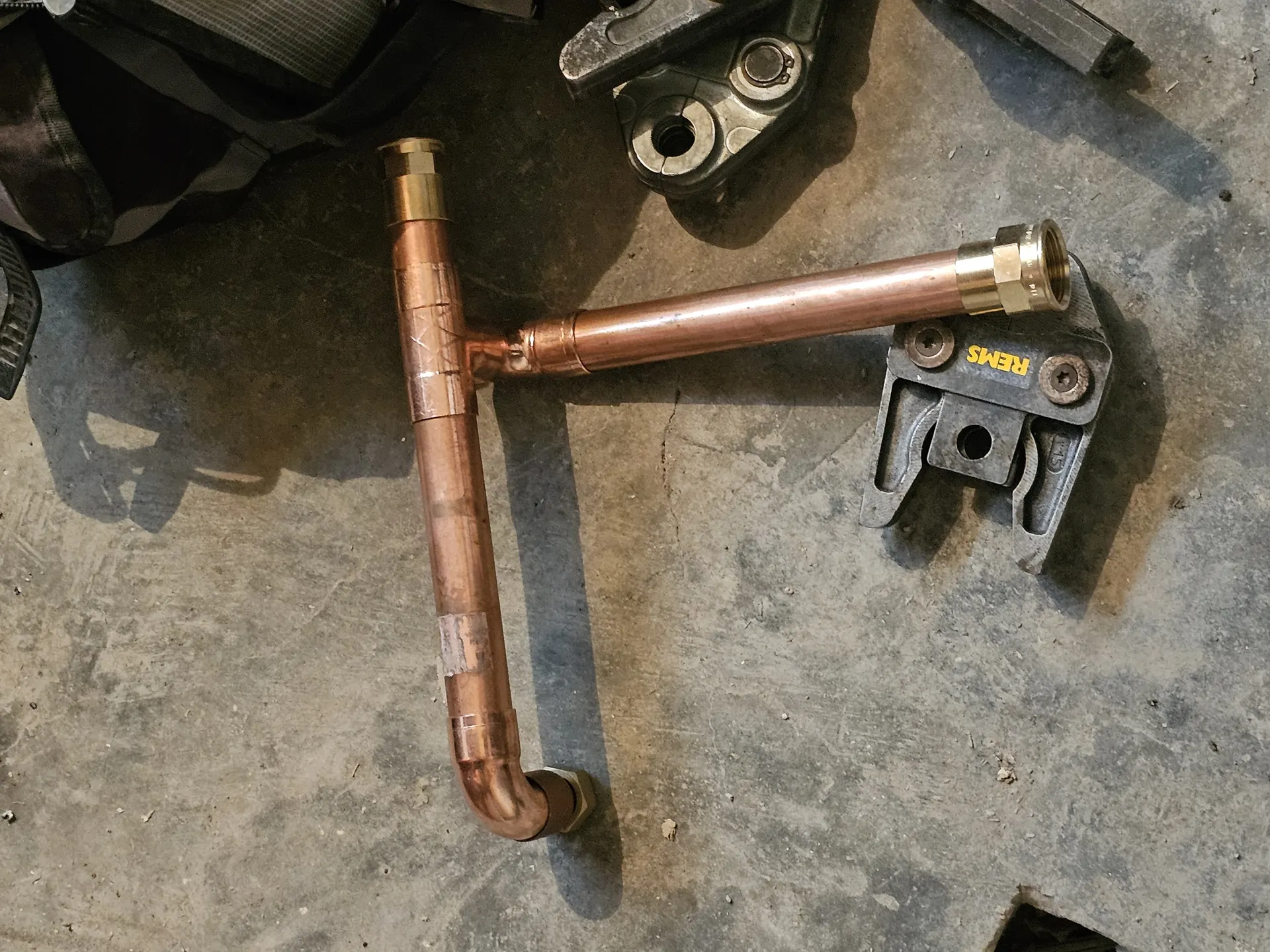 A képen egy rézcső látható, amely az épületgépészethez szükséges alkatrészek közé tartozik, és a HydroArt Kft. Öskü városában végzett munkáira utal. A cső ívelt formában van, és különböző szerelési elemekkel, például csavarokkal és rögzítőkkel van körülvéve, amelyek a telepítési folyamat részei. A háttérben látható egy szerszámos táska, ami a szakmai környezetet tükrözi.