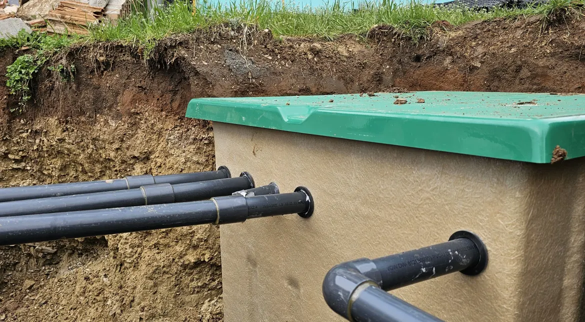 Az Ösküben található HydroArt Kft. épületgépészeti projektje során készült kép, amelyen egy zöld fedelű tartály látható, amelyhez fekete PVC csövek csatlakoznak. A tartály egy ásott gödörben helyezkedik el, a háttérben látható a föld kiemelkedése és a növényzet. Az épületgépészet területén végzett munkák precizitását és szakértelmét tükrözi ez a részlet.