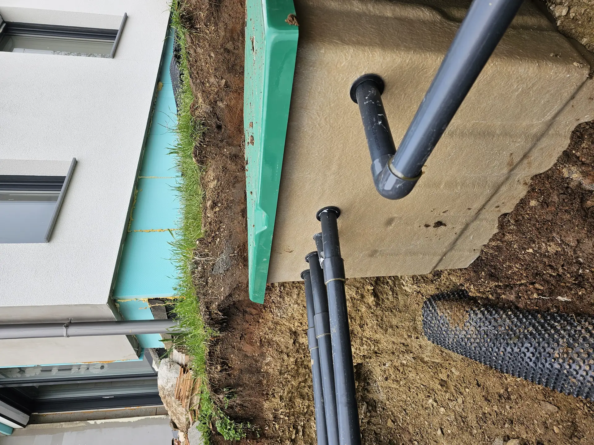 Az Ösküben található HydroArt Kft. épületgépészeti projektje során készült képen egy új vízelvezető rendszer látható. A zöld színű burkolat alatt fekvő szürke beton alap és a fekete PVC csövek jól megfigyelhetők. A munkaterület rendezett, a fű is látható a talaj mellett, ami a projekt precizitását tükrözi.